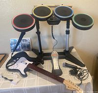 Photo showing full drum kit with four colored pads mounted on stands, bass pedal, Fender style guitar controller, wired microphone, and two Wii game cases (Rock Band 2 and The Beatles Rock Band).