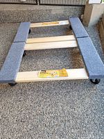 Photo showing one furniture dolly, wooden frame with grey padded strips along two longer sides, 3 caster wheels underneath