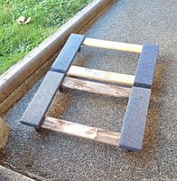 Top view of weathered wood furniture dolly with gray padding on edges, laid outside on pavement next to grass.