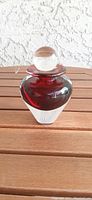 Front view of the red and clear blown glass perfume bottle with clear spherical stopper, placed on wooden surface with textured white background.