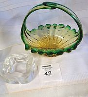 Bullicante green and amber glass basket bowl with handle, alongside clear glass ashtray, displayed on white textured cloth.
