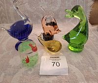 Five unique glass animal paperweights displayed on a textured cloth background including a blue swan, pink cat, yellow whale, green crocodile, and clear dome with red fish.
