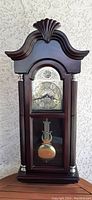 Front view of dark wood mantle style wall clock showing ornate curved top with shell design, glass door revealing pendulum and clock face with silver tone and black numbers.
