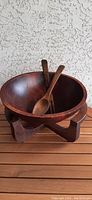 Wooden salad bowl set on wooden stand with two wooden utensils inside, photographed from front angle against stucco wall.