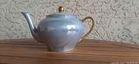 Side view of a small light blue teapot with gold handle and gold knob on lid, positioned on a wooden surface.