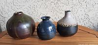 Three pottery vases displayed on a wooden surface against a textured wall, showing their different colors and shapes