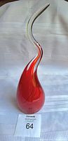 Full view of the red and clear glass drop sculpture showing teardrop base and curved upward neck on white fabric background.