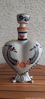Front view of the heart-shaped white pottery tequila bottle with blue and orange floral designs and stopper.