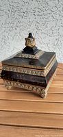 Side view of decorative treasure box showing ornate gold-toned finial on the lid, black base with gold mottling and decorative gold trim on edges and feet.