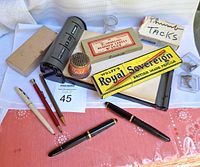 Photo showing wide view of vintage office desk items on pink cloth including pencils, pens, grey date planner, boxed pencils, boxes of gum rolls and thumb tacks.