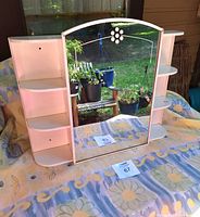Front view of vintage enamelled metal medicine cabinet showing curved top mirror with floral etching and open shelving units on both sides.