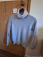 Front view of light grey thin knit angora sweater hanging on a wooden door. Shows classic turtleneck and sleeves with fuzzy texture.