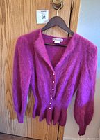 Front view of vintage purple angora wool sweater hung on a hanger against a wooden door. Shows full length and texture of thick knit and fuzzy angora fibers. Six faux pearl buttons visible.