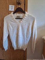 Front view of a vintage off-white angora wool sweater hanging on a hanger on a door. Thin knit with bow string at neckline visible.