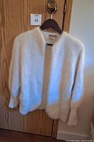 Front view of vintage angora wool sweater jacket hanging on a wooden hanger against a wooden door, showing its milky white thick fuzzy knit, open front, and two small pockets near the hem.