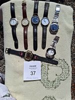 Top view of seven wrist watches arranged on cloth, showing watch faces and straps in various materials and styles.