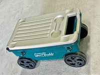 Side view of green and tan Ames Lawn Buddy garden cart with four black wheels and handle on tan lid.