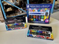 Photo showing three acrylic paint sets: one TBC The Best Crafts 28-color paint and brush set, and two Winsor 12-color acrylic paint sets with brushes.