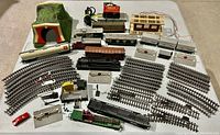 Wide view of assorted model train items including locomotive engines, freight and passenger cars, tracks, switches, buildings, and a mountain tunnel arranged on a flat surface.