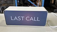 Blue message light box with white text "LAST CALL" on front face, rectangular shape, placed on a table with industrial shelving in the background.