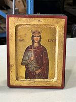 Front view of religious icon depicting a crowned Saint Barbara holding a cross and palm branch, set in a recessed gold-toned background within a wooden plaque frame.