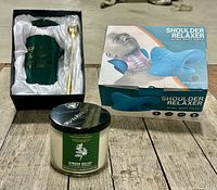 Lot contents showing green ceramic mug with gold spoon in satin box, aromatherapy stress relief candle, and blue shoulder relaxer in original box on wood floor.