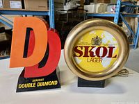 Side-by-side photo of the Double Diamond Draught light with 3D red and orange DD letters and Skol Lager round sign with a gold rim and yellow face
