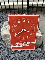 Front view of vintage Coca-Cola metal wall clock on gravel with rock background showing red clock face, white numbers, hands, and Coca-Cola logo.