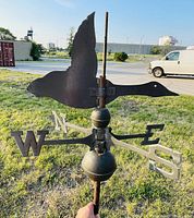Weather vane at an angle showing flying goose metal silhouette and directional letters mounted on metal rod and wood stake outdoors on grass with vehicles in background.