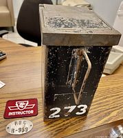 Side angle of vintage heavy cast coin collection box with visible number 273, metal latch, TTC Instructor patch and metal plaque on wood surface.
