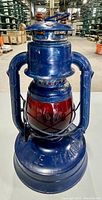 Front view of the vintage blue metal lantern showing 'Little Wizard' embossed on the base and the red glass globe protected by metal crossbars.