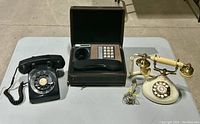 Lot of three vintage telephones: a black rotary dial phone, a push-button 'Decotel' phone in a hard case, and a cream brass rotary phone with ornate handset.