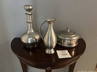 Three-piece mid-century modern pewter set displayed on a wooden half-moon table including a decanter with stopper, a pitcher, and a lidded covered dish.