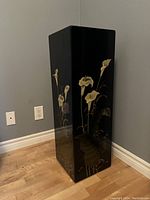 Black lacquered rectangular pedestal shown from angled side view, highlighting floral design and glossy finish.