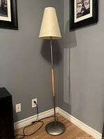 Postmodern floor lamp with beige fabric conical shade, circular metal base, and wooden accent on the metal pole, standing in a corner against gray walls.