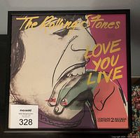 Framed front cover of The Rolling Stones "LOVE YOU LIVE" double album with distinctive pop art style by Andy Warhol. The cover features the title text and a stylized side profile face with fingers near the mouth colored with nails painted red.