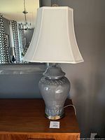 Front photo of vintage ceramic ginger jar lamp showing shiny gray ceramic body with brass base and white fabric shade on a wooden surface.