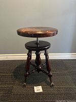 Full front view of vintage adjustable piano stool showing round wooden seat, height adjustment screw, four turned legs, and claw feet with glass balls.