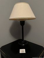 Photo of the full vintage postmodern table lamp showing the cream fabric lampshade and black glossy base placed on a black surface against a gray wall.