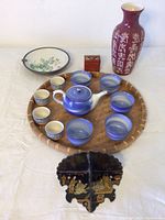Blue ceramic teapot and matching cups arranged on a round bamboo tray with additional ceramics and decor items behind