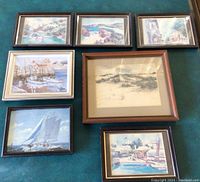 Top down view showing seven framed watercolor and one sketch painting arranged on green surface, depicting boats and coastal scenes