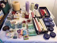 Wide view of large assortment of candles including pillars, tapers in boxes, birthday candles, and candle holders in blue glass and metal.