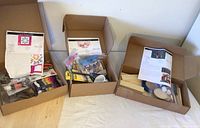 Three open craft kits shown side by side with instruction sheets and various components inside cardboard boxes.