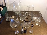 Clear glass vases and pitchers in various shapes and sizes on table including large snifter, pitchers with silver rims, and square vases.