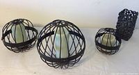 Front view of three black iron caged sphere candle holders of varying sizes with white LED candles inside, alongside black rectangular decorative candle holder.