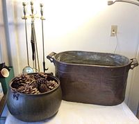 Two large copper kettles side by side on a white surface, one filled with pinecones, with a brass fireplace tool set standing next to them.