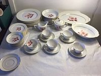 Front view of the full Mikado Prima Donna china set laid out on a white cloth including various plates, bowls, cups, saucers, sugar bowl, and serving pieces with pink and red rose design and gold trim.