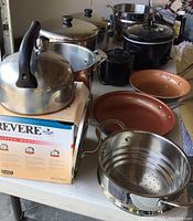 View of assortment of cookware items including stainless steel kettle, pots, steamer basket, and ceramic frying pans on table.