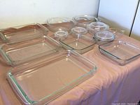 Multiple Pyrex glass baking dishes including rectangular and round shapes, some with lids, arranged on pink fabric.