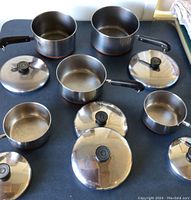 Overview of five stainless steel pots with copper bottoms and black plastic handles, arranged with their corresponding lids on a table.